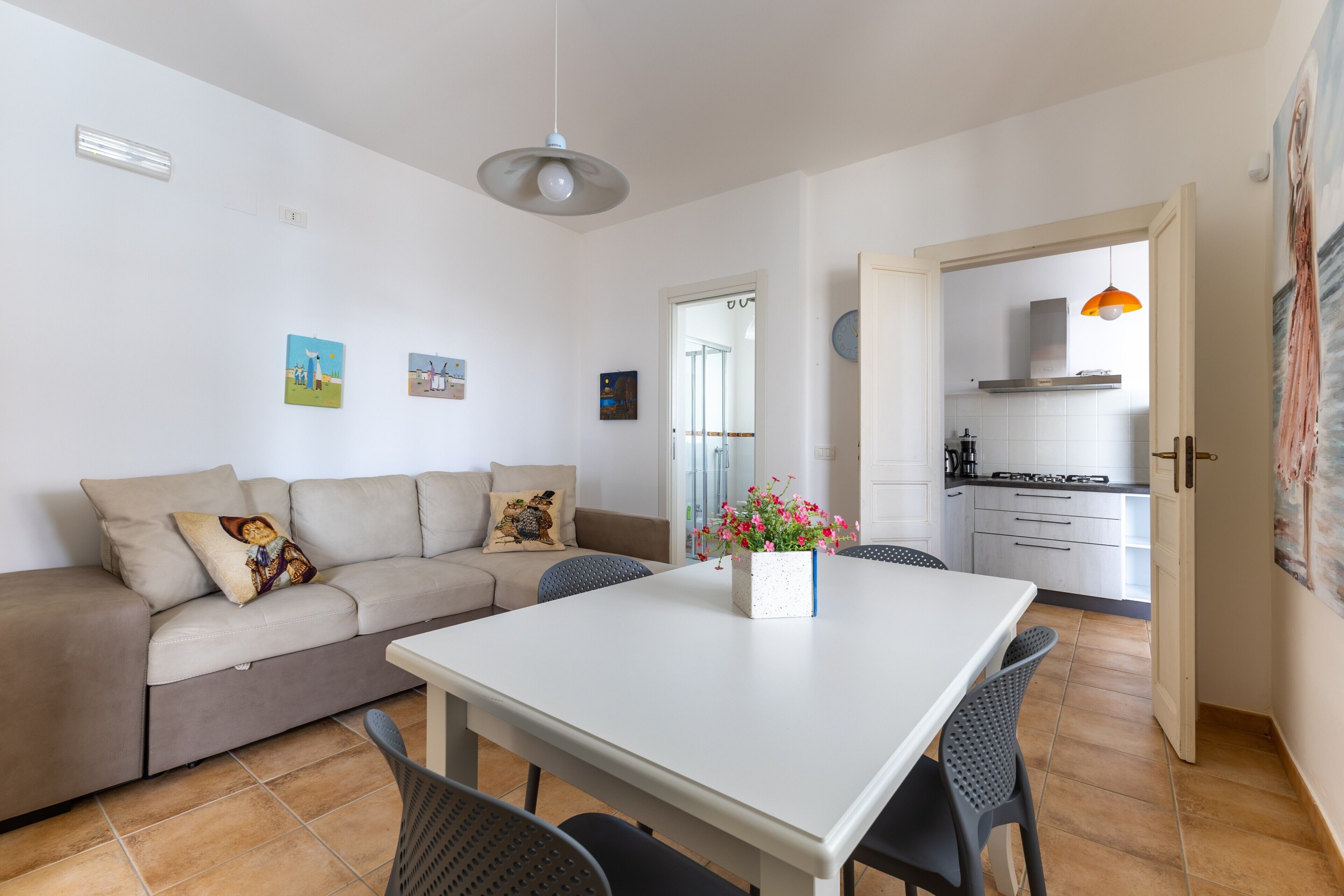 A modern living room with a sofa, a dining table, and chairs. There is a kitchen visible through an open doorway.