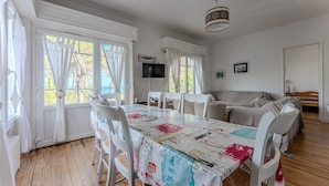 Un coin repas avec une table recouverte d’une nappe colorée, des chaises blanches et un téléviseur fixé au mur.