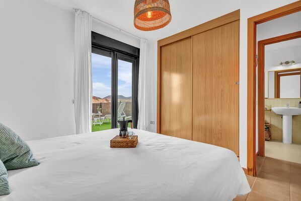 Ein Schlafzimmer mit Bett, einem Holzschrank, einem Badezimmer mit Waschbecken und Blick ins Freie.