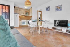 Un salon moderne avec un canapé, une table à manger et des chaises, un téléviseur, et une cuisine en arrière-plan.