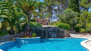 Una piscina con caída de agua, rodeada de abundante vegetación y un muro de piedra.