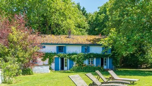 Een huis met een pannendak, blauwe luiken en een tuin met ligbedden.