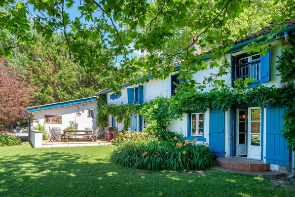 Una casa de dos pisos con un porche cubierto, un pequeño jardín con flores y una mesa y sillas de madera.