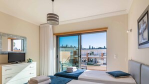 Ein Hotelzimmer mit Bett, Fernseher, Schminktisch und Balkon mit Blick auf Gebäude.