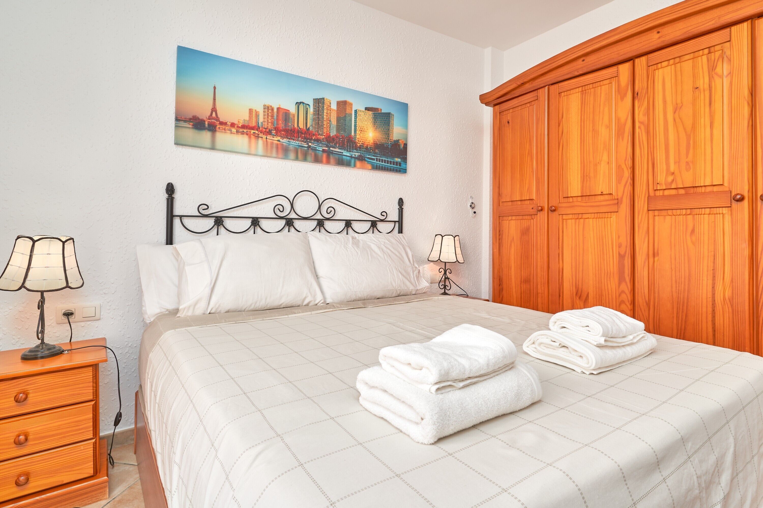 A hotel room with a bed, two bedside lamps, a wooden wardrobe, a cityscape painting on the wall, and a nightstand with a lamp.