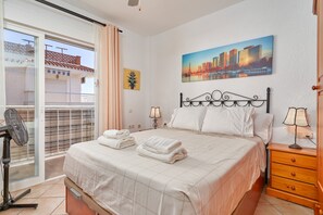 Une chambre d’hôtel avec un lit, une table de chevet, une lampe, une fenêtre avec des rideaux et un tableau représentant un paysage urbain accroché au mur.