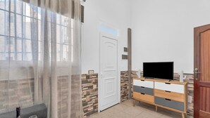 Una habitación con una televisión sobre un mueble de madera, una puerta blanca, una ventana con cortinas translúcidas y un suelo de baldosas.