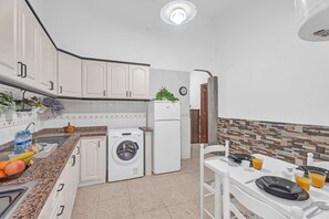 Une cuisine avec des meubles blancs, un lave-linge, un réfrigérateur, une table à manger dressée avec des assiettes, des verres et une horloge au mur.