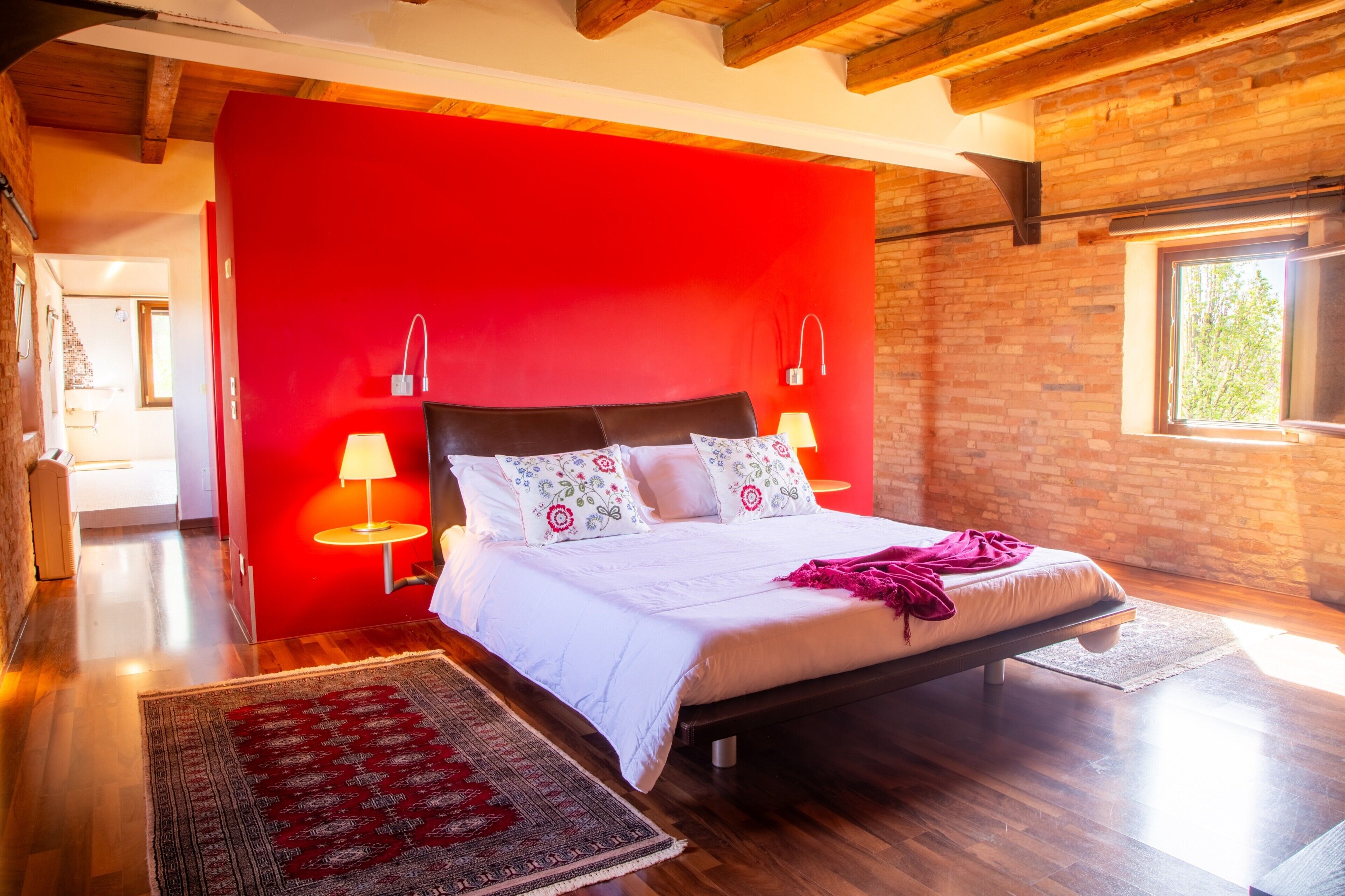 Un dormitorio con una pared de acento roja, un cabecero de madera, dos lámparas de noche y un ventanal grande.
