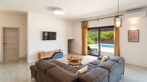 Uma sala de estar moderna com um sofá seccional, uma mesa de centro, uma televisão de tela plana e uma porta de vidro deslizante que dá para uma área de piscina.
