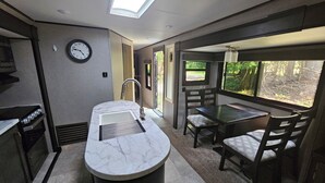 A compact kitchen with a marble countertop, a sink, a clock, and a window overlooking a wooded area.