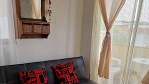 A black leather sofa with red patterned cushions, a wooden wall-mounted shelf with hooks, a ceiling fan, and sheer curtains. - Cabañas Aquila D'Arroscia (Caldera)