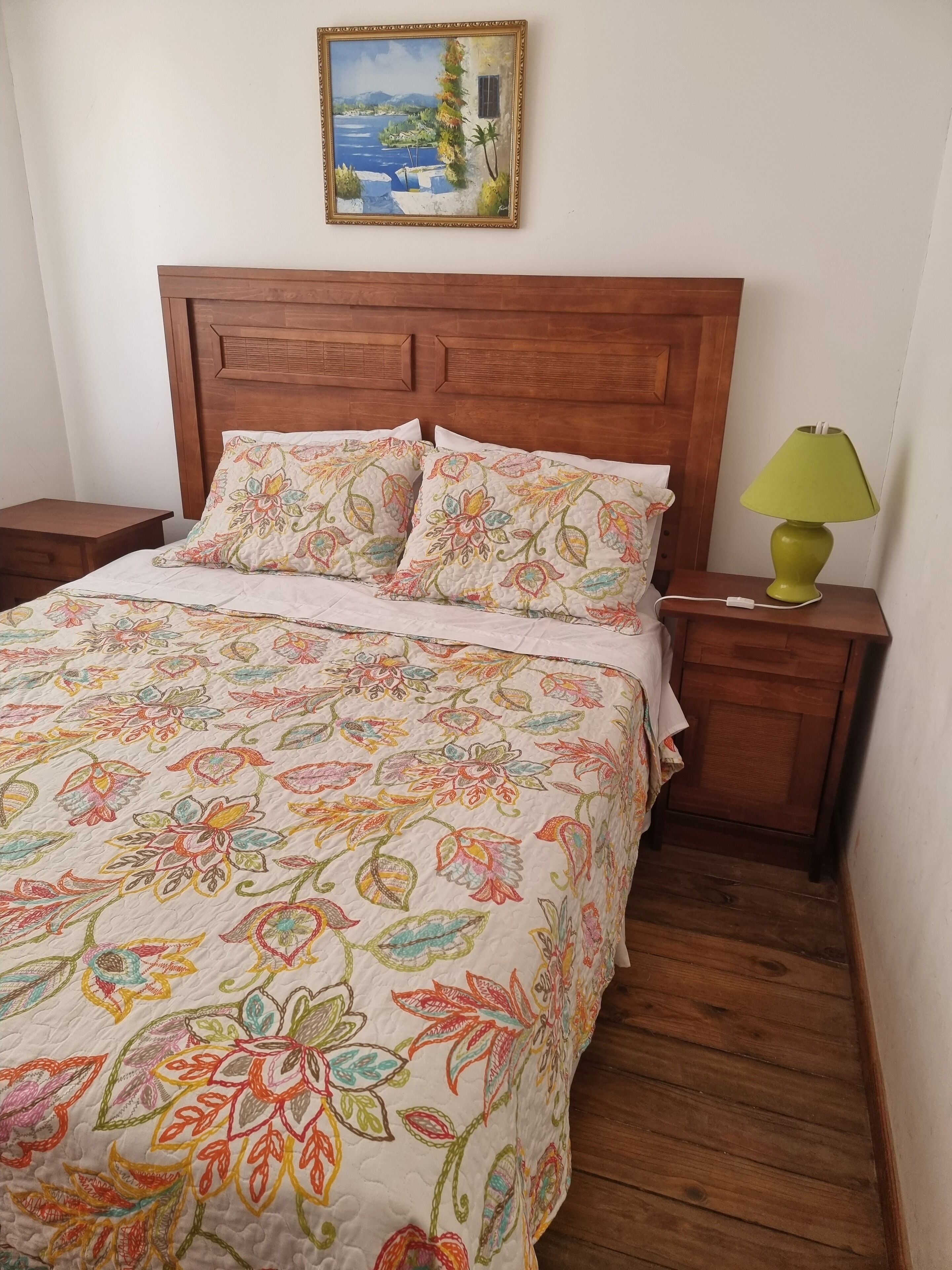 A bedroom with a wooden headboard, floral bedspread, two matching pillows, a green table lamp, and a painting of a landscape on the wall.