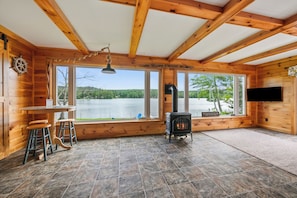 Eine Hütte mit großem Fenster, Kamin, Holztisch mit Hockern und einem an der Wand befestigten Fernseher.