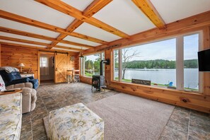 A cabin with a fireplace, a sofa, a dining area, and a large window overlooking a lake.