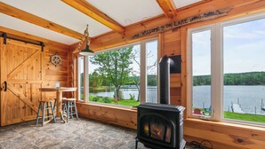 Ein Holzblockhaus-Interieur mit Kamin, großen Fenstern, einem Esstisch mit Hocker und Blick auf einen See.