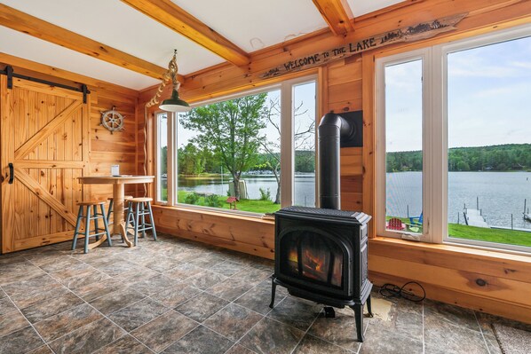 A wooden cabin interior with a fireplace, large windows, a dining table with stools, and a view of a lake.