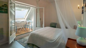 A bedroom with a bed, a nightstand, a lamp, a chair, and a view through an open window to an outdoor area with a table and chairs.