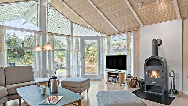 A modern living room with a fireplace, comfortable seating, a television, and large windows offering a view of the outdoors. - Beautiful home in ålbæk with sauna (Ålbæk)