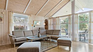 A cozy living room with a sectional sofa, a small table, and an ottoman. There are large windows with curtains, a lamp, and a framed picture on the wall. - Beautiful home in ålbæk with sauna (Ålbæk)