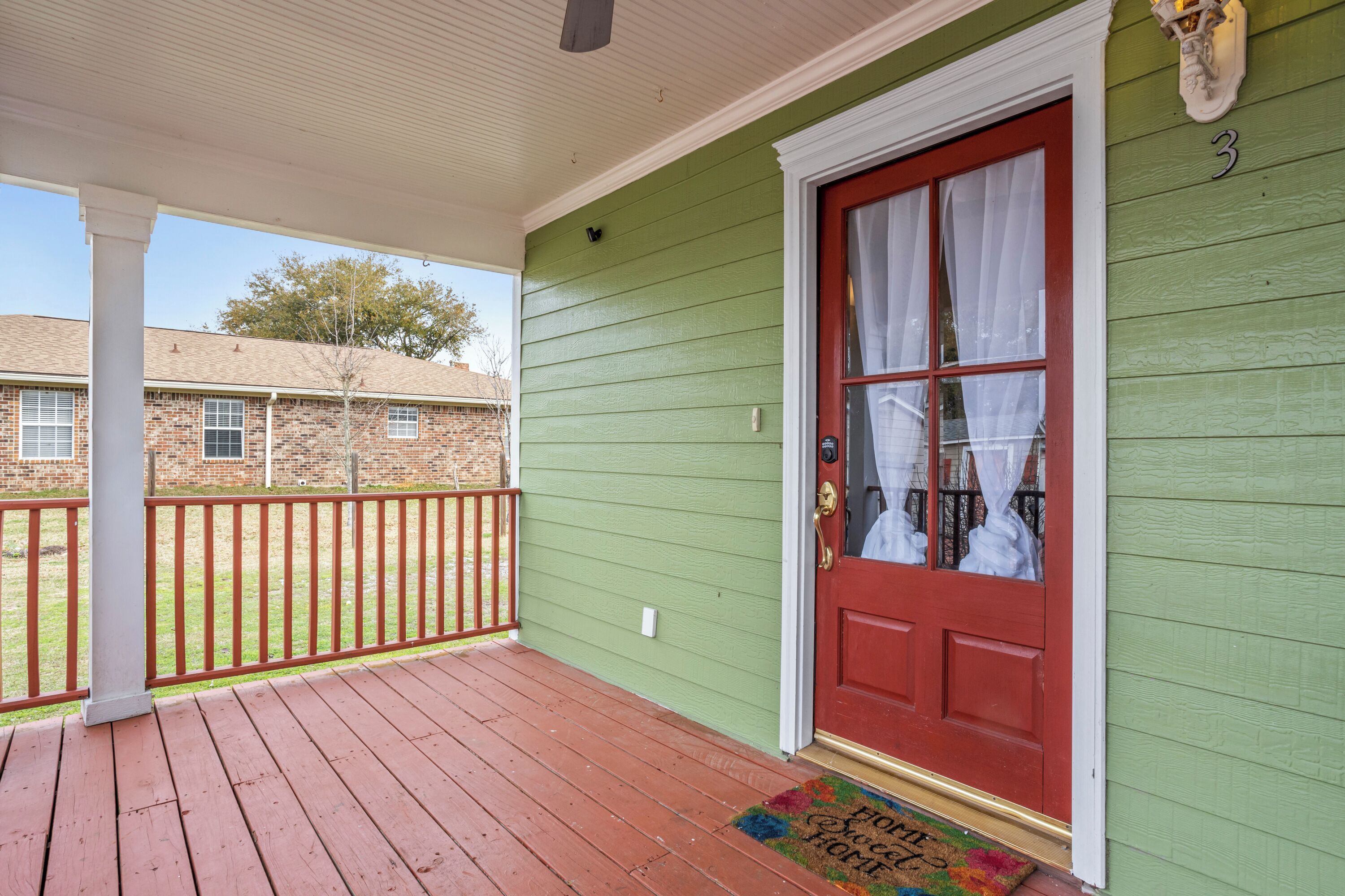 A green house with a red door, a porch with a red railing, and a brick building in the background.
