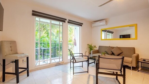A spacious living room with a sofa, chairs, and a coffee table. Large windows provide natural light and a view of greenery outside. - Jardín Departamento Privado 2 Habitaciones Cancún (Cancún)