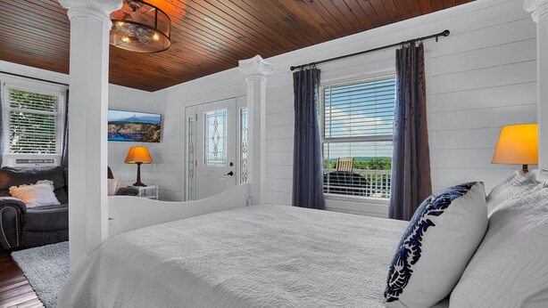 A bedroom with a large bed, a nightstand with a lamp, a window with curtains, and a view of greenery outside. - Romantic Golden Valley Cottage | View of Amish Countryside + Walk to Amish Eats (Sugarcreek)
