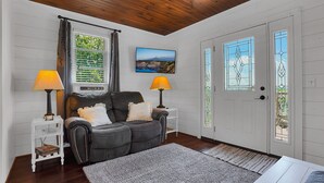 A cozy room with a dark leather sofa, two side tables with lamps, a framed picture on the wall, and a white door with stained glass windows. - Romantic Golden Valley Cottage | View of Amish Countryside + Walk to Amish Eats (Sugarcreek)