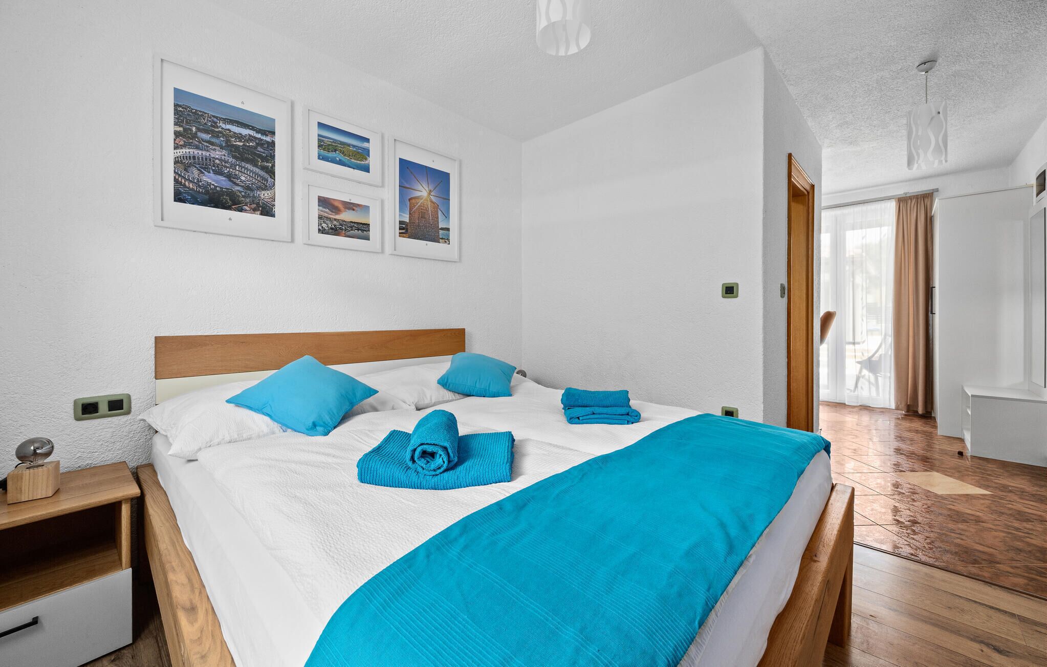 Una cama bien hecha con ropa de cama azul, un cabecero de madera y una mesita de noche con una lámpara. Hay tres cuadros enmarcados en la pared sobre la cama.