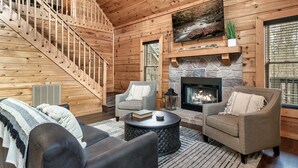 A cozy cabin interior with a stone fireplace, a staircase, and two armchairs.