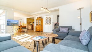 Ein Wohnzimmer mit grauem Sofa, Couchtisch und Fernseher. Im Hintergrund befindet sich ein Küchenbereich mit Holzschränken und einem Herd.