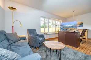 Un salon moderne avec un canapé gris, un fauteuil bleu, une table basse en bois et un téléviseur diffusant une scène de plage.