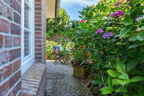 Un mur extérieur en briques avec une fenêtre, un chemin pavé de pierres, un jardinier en bois et diverses plantes à fleurs.