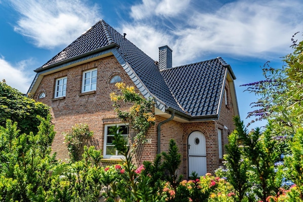 Une maison à deux étages, construite en briques, avec un toit à forte pente, une cheminée et un petit jardin où poussent diverses plantes et fleurs.