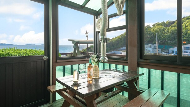 Una mesa de madera con una botella y vasos, una lámpara colgante y vistas al mar y edificios.