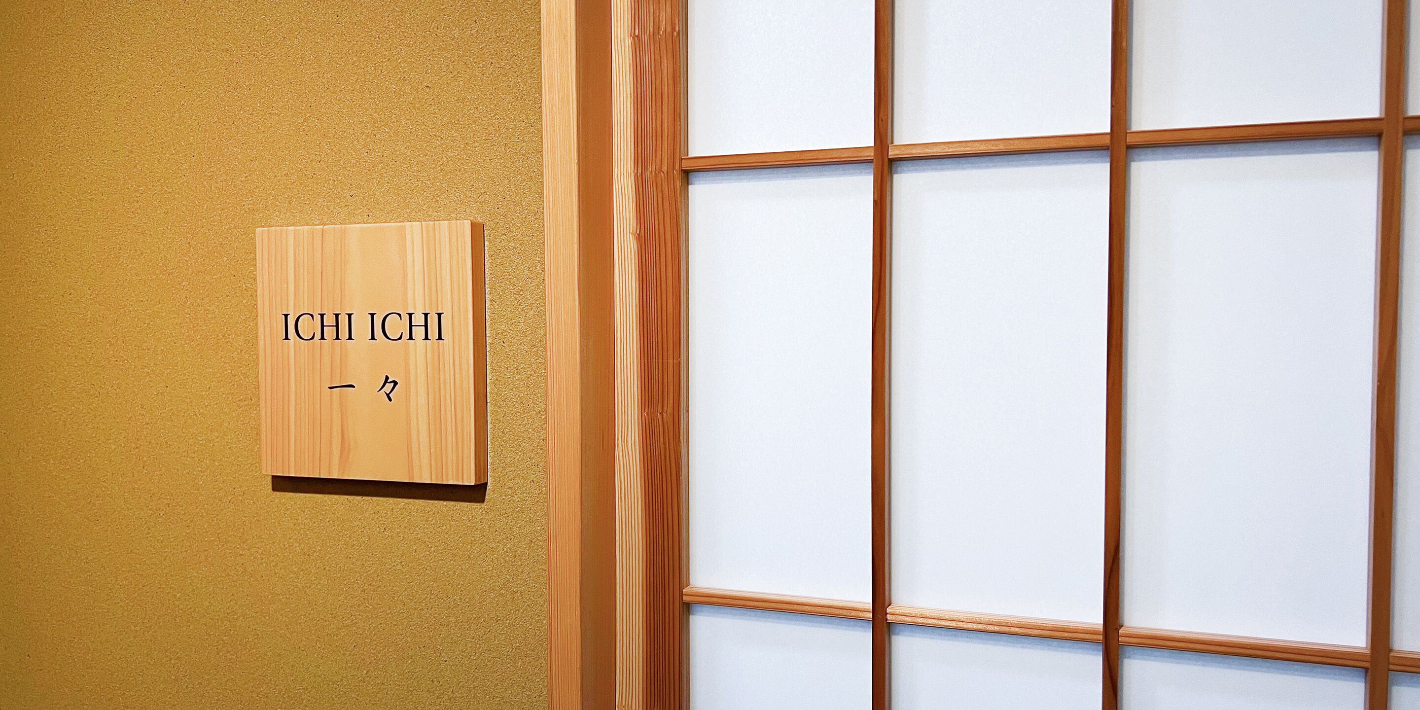 Une enseigne en bois, ornée de caractères japonais, est fixée à un mur, à côté d’une porte coulissante traditionnelle à motifs géométriques.