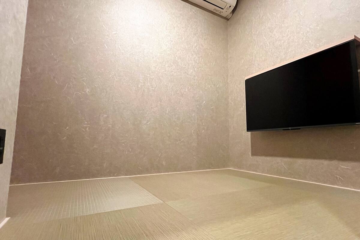 Una habitación con una televisión de pantalla plana montada en la pared, un revestimiento beige texturizado y piso de baldosas.