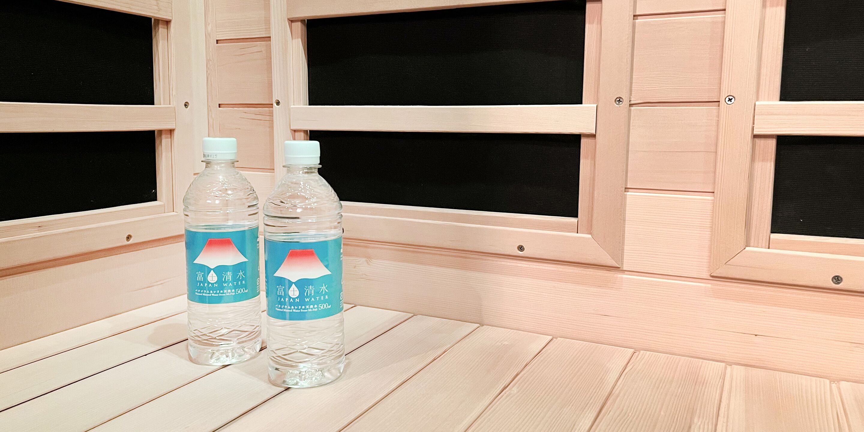 Dos botellas de agua japonesa sobre un suelo de madera con fondo negro y paneles de madera.