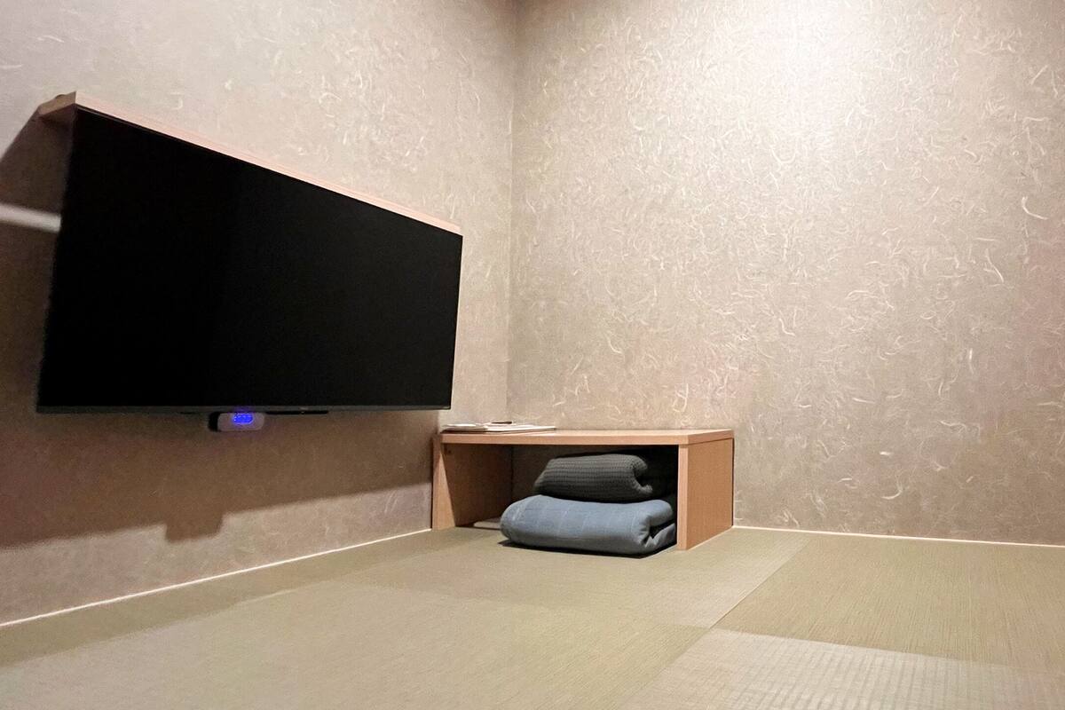Una habitación con una televisión de pantalla plana en la pared, una pequeña mesa de madera y una zona de estar con cojín.