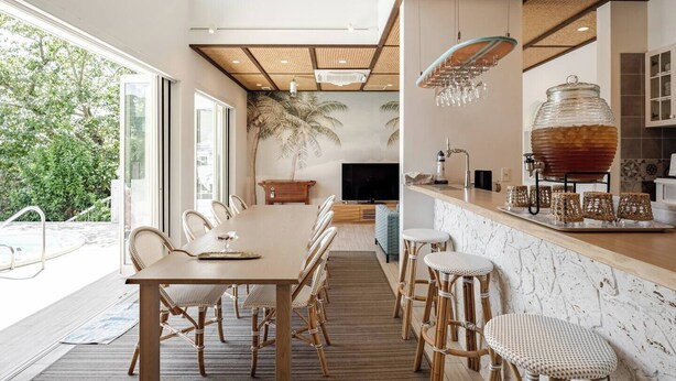 Una cocina moderna con una isla grande, un comedor con una mesa larga y sillas de mimbre, y un televisor grande al fondo.