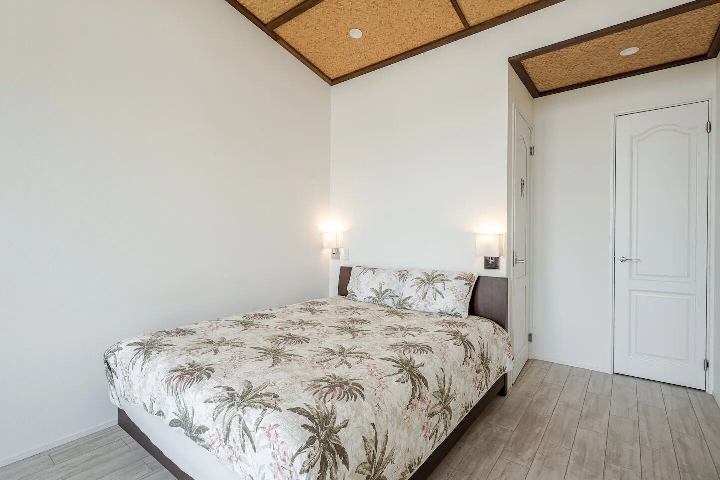 A bedroom with a bed, two wall-mounted lights, a door, and a ceiling with wooden beams.