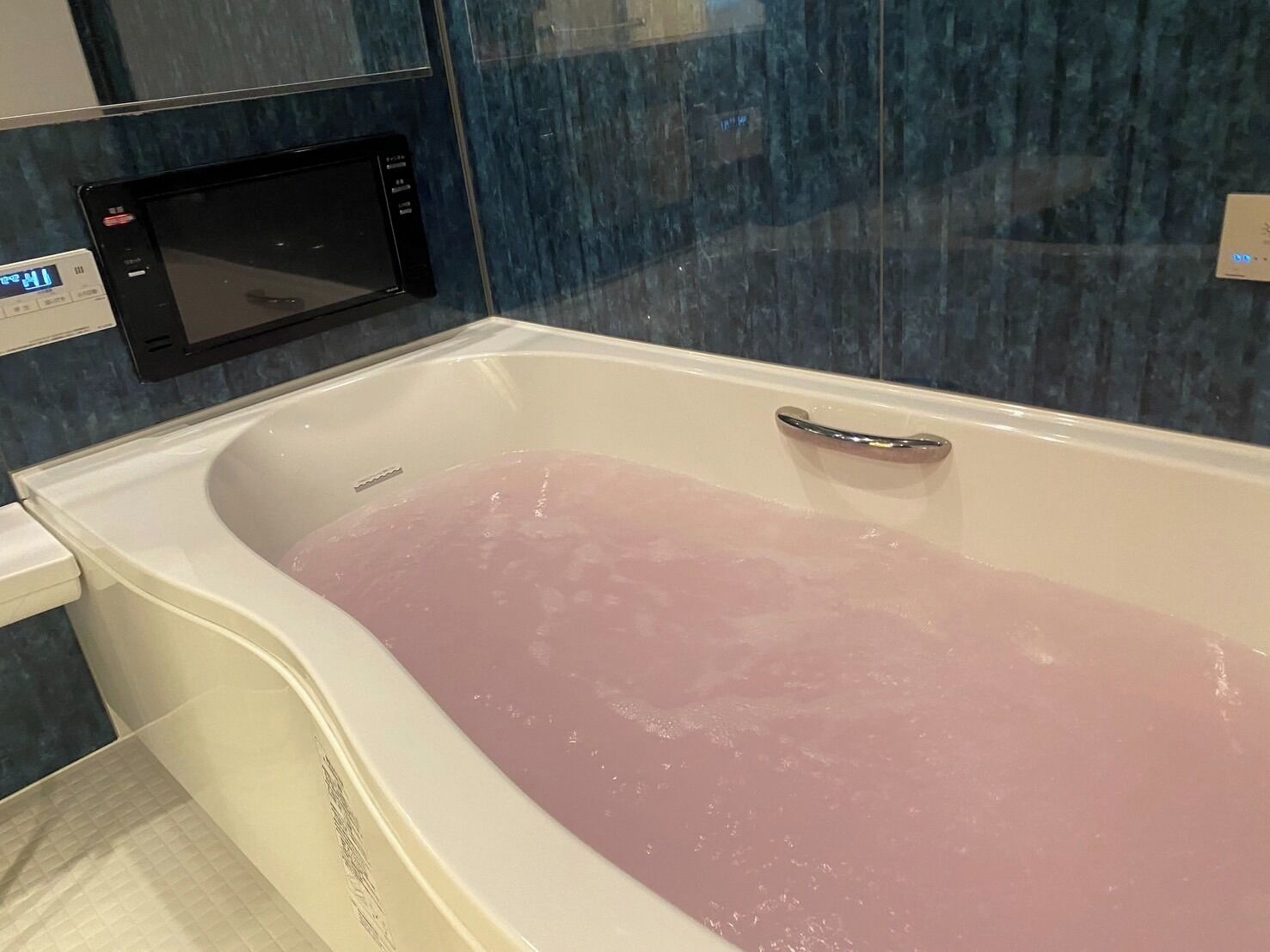 Une baignoire remplie d’eau rose, une horloge numérique et un panneau de commande au mur.