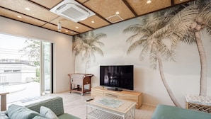 Una sala de estar moderna con un televisor de pantalla plana grande, una mesa de centro de madera y un sofá. Se observa un mural de palmeras en la pared y un techo con iluminación empotrada.