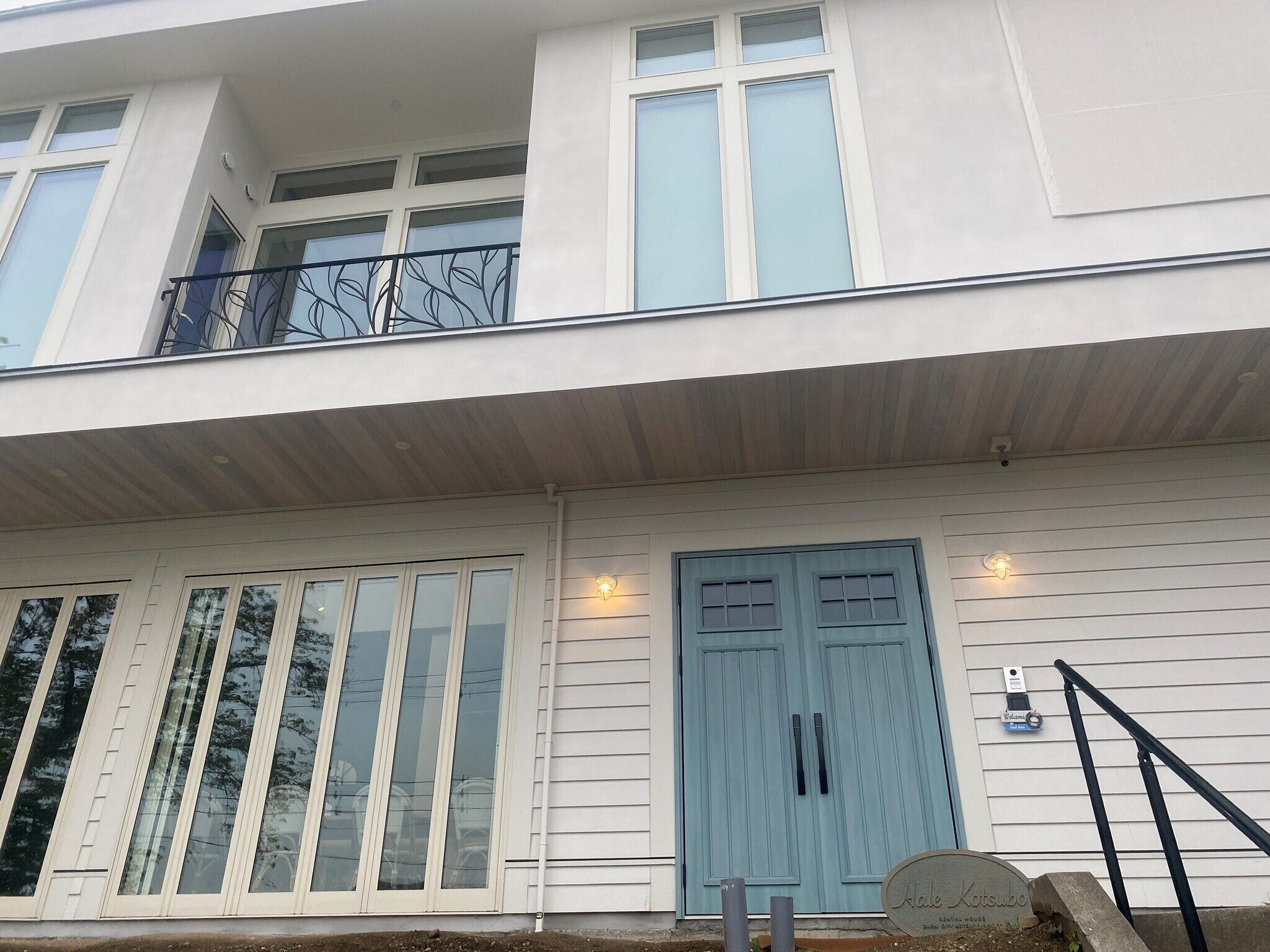 A two-story building with a light blue door, large windows, and a balcony with a metal railing.