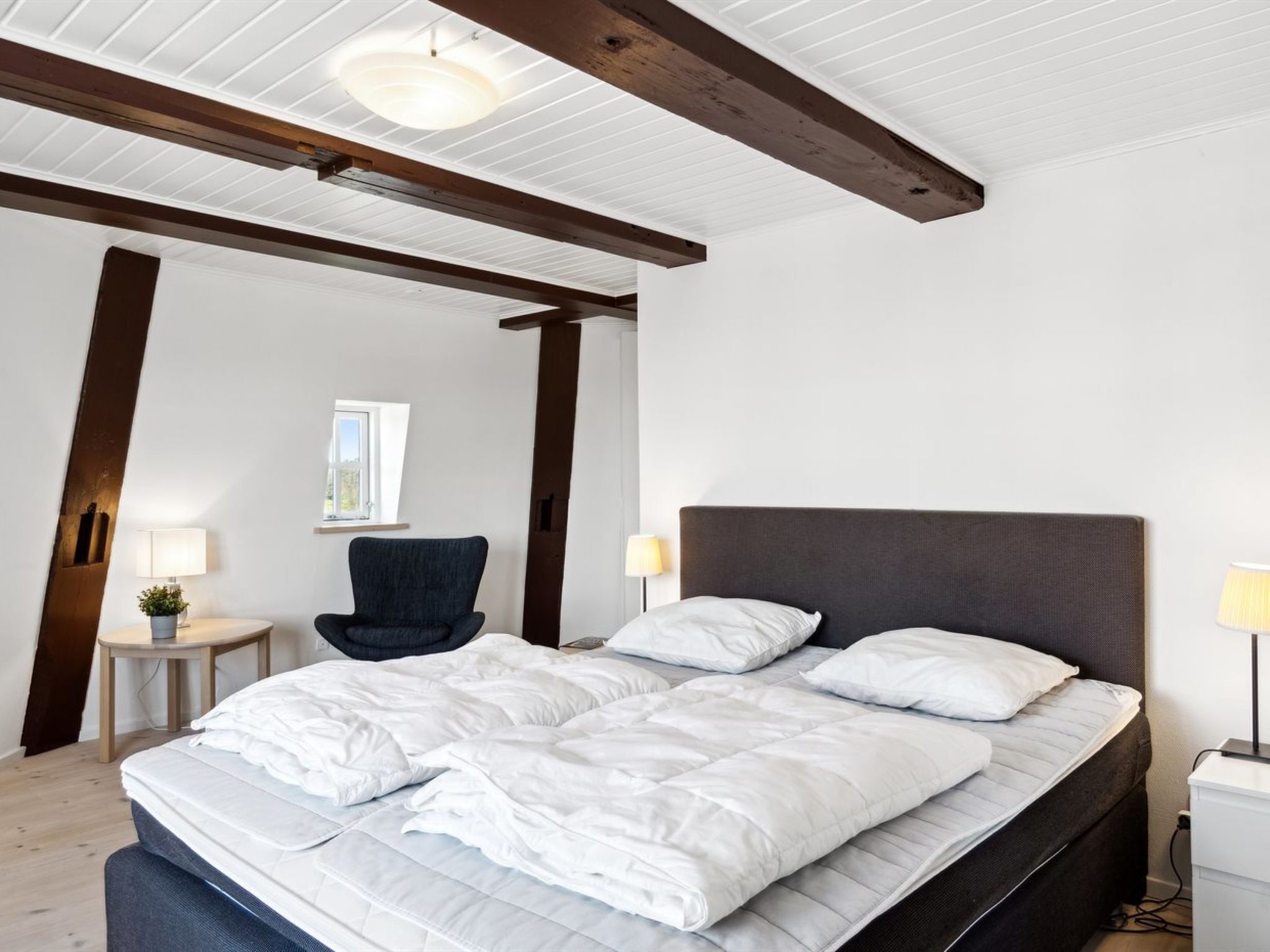 A bedroom with a large bed, wooden beams, a chair, and a small table.