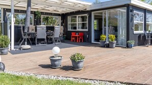 Un patio exterior moderno con terraza de madera, zona de estar cubierta con sillas y una mesa, y plantas en macetas.
