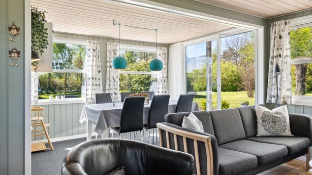 Una sala de estar acogedora con un sofá gris, un sillón de cuero negro y un comedor con mesa y sillas. Amplias ventanas ofrecen vistas a un jardín.