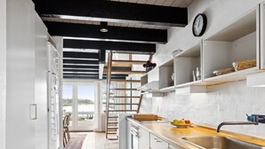 Una cocina moderna con armarios blancos, encimera de madera y pileta de acero inoxidable. Hay un ventanal con vista al exterior y una escalera que lleva a un altillo.