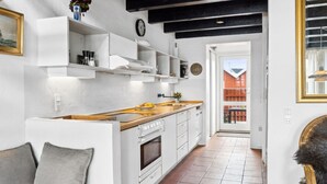 Una cocina moderna con armarios blancos, una encimera de madera y un horno empotrado. Cuenta con una zona de comedor con una mesa blanca y cojines grises. La cocina tiene un techo negro y una ventana con vistas a un edificio rojo.