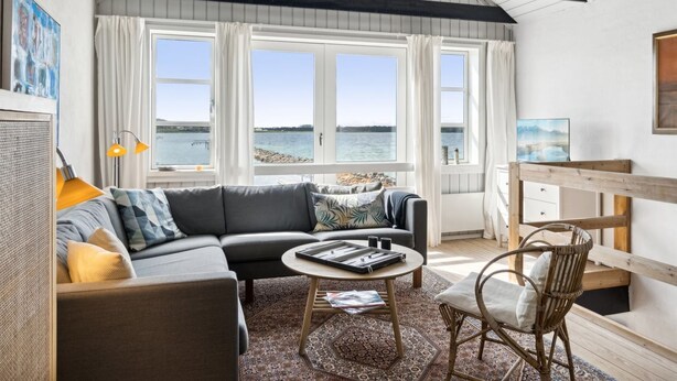 Un salón acogedor con sofá, mesa de centro y sillón de mimbre, amplias ventanas con vista al agua y una viga de techo de madera.